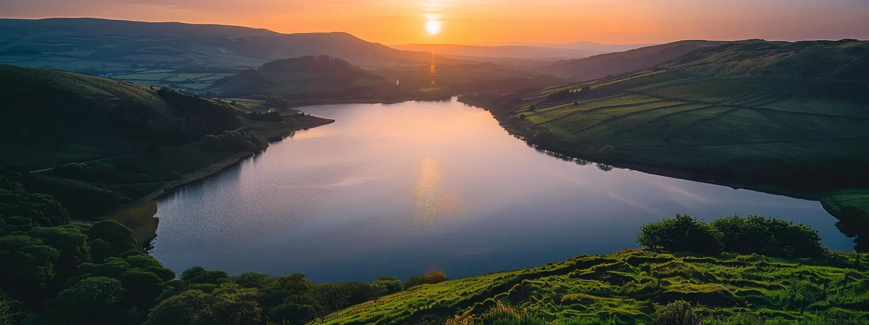 a breathtaking sunset illuminates a tranquil lake, with vibrant hues of orange and pink reflecting off the glassy water, surrounded by lush green hills under a clear sky.